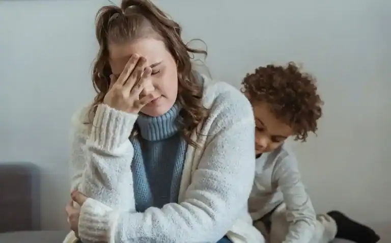 Una madre con cara de angustia se tapa la cara con la mano mientras un niño pequeño la observa, ilustrando el desborde emocional y el estrés parental.